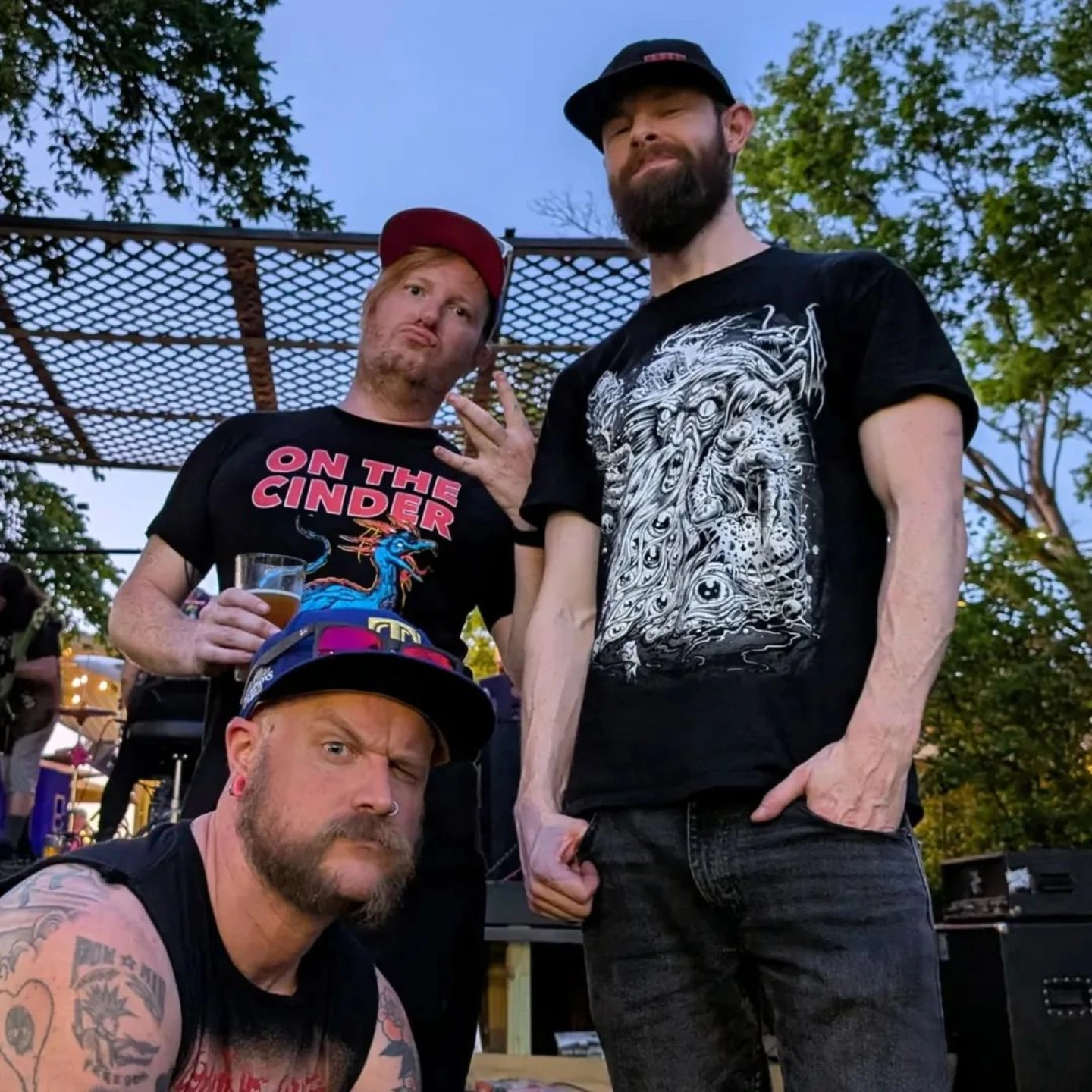 Three men standing outdoors, two wearing black t-shirts with graphic designs, one holding a drink.