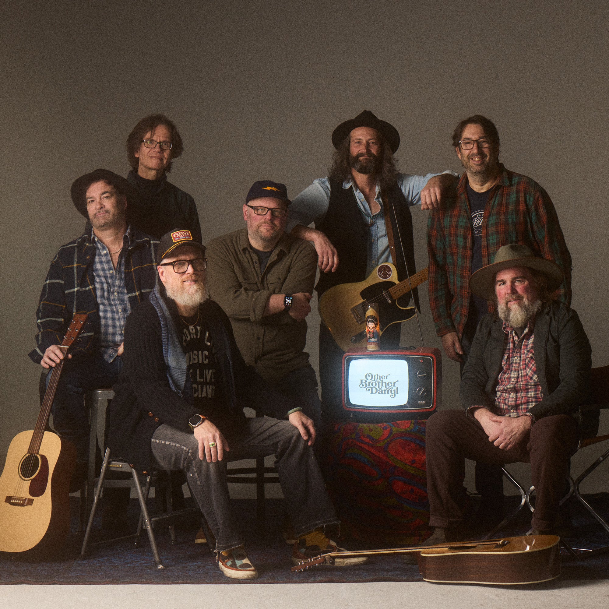 Image shows OBD - a new ONE on Stage band. Seven men are looking at the camera. Some or standing and some are sitting. There are three guitars and a TV placed around them.