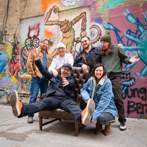 Image of Amateur - a ONE on Stage band. Six band members infront a graffiti printed building. One person sits on a brown chair. 