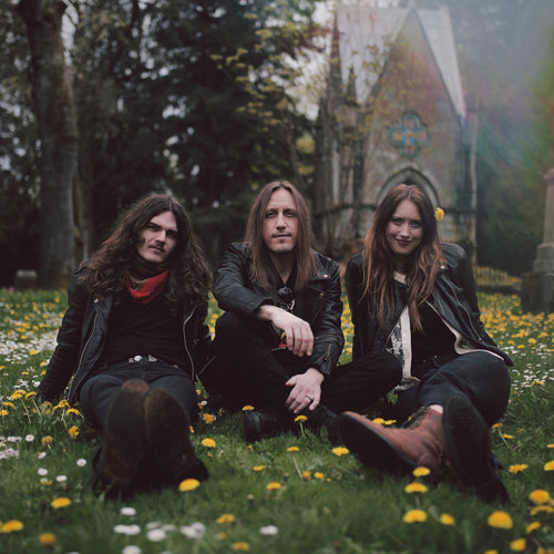 Image of Hippie Death Cult - a new ONE On Stage band. Image shows three people (band members) sitting in a bed of grass dotted by dandelions. All band members are wearing black. A church-looking stone building and trees are behind them.