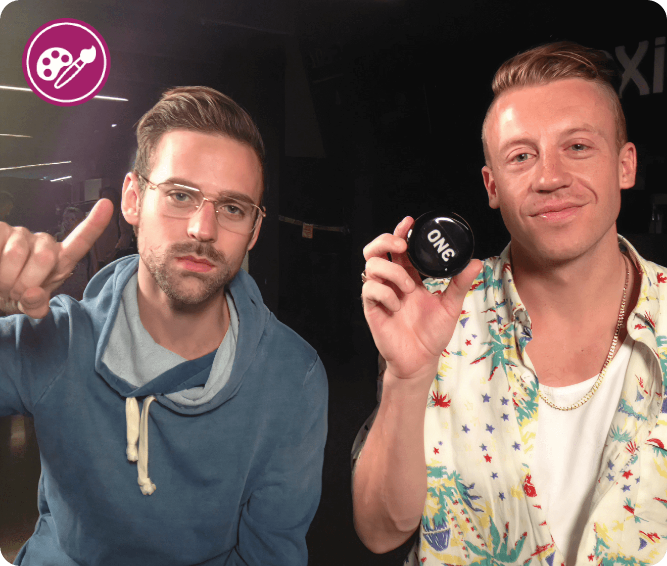 Two men posing for a photo, one pointing and the other holding a small round object.